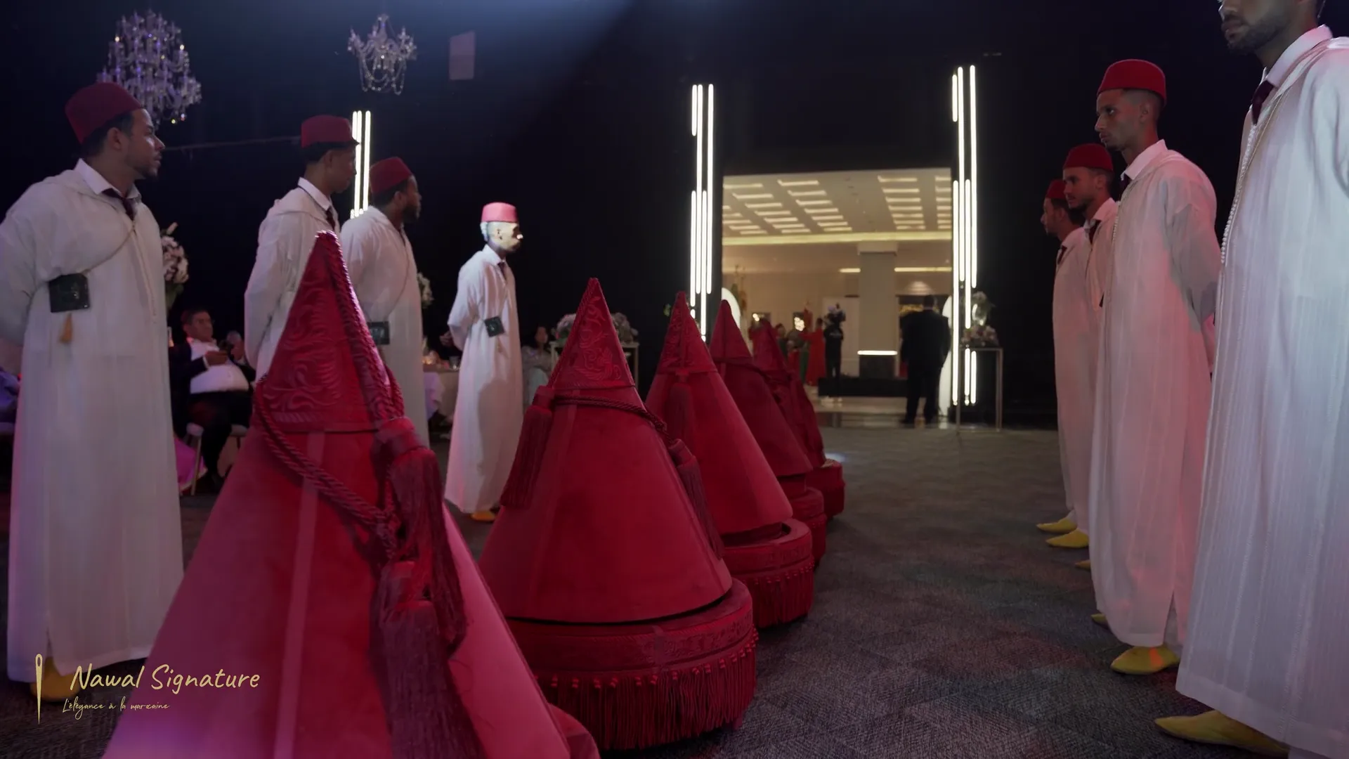 Rangée de Tyafer en velours rouge portés par des porteurs en djellaba blanche et fez, dans une salle de réception